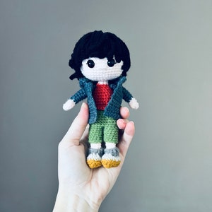 May include: A crocheted doll with black hair, a blue jacket, a red shirt, green pants, and yellow shoes.