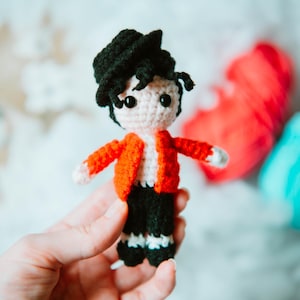 Puede incluir: Muñeca de ganchillo hecha a mano de una persona con sombrero negro, chaqueta roja, pantalón negro y calcetines blancos. La muñeca tiene la cara blanca y el pelo negro. La muñeca se sostiene en una mano, con un objeto rojo y azul en el fondo.