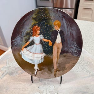 May include: Decorative plate featuring a painted scene of a boy and girl dancing in front of a Christmas tree. The girl wears a light blue dress with a red sash. The boy is in a black jacket and tan pants. The plate is displayed on a black stand.