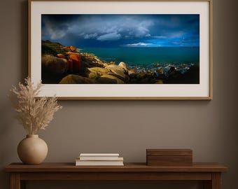 May include: A framed landscape photograph featuring a coastal scene with large rocks, turquoise water, and a dramatic sky with dark clouds. The photograph is displayed on a wooden console table with a vase of dried flowers and a small wooden box.