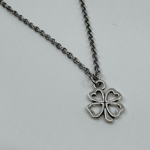 Puede incluir: Collar plateado con un colgante de trébol de cuatro hojas. El colgante presenta un diseño calado detallado, y la cadena es de estilo eslabones delicados. El collar se muestra sobre un fondo blanco liso.