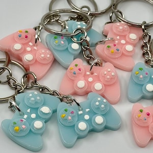 Könnte beinhalten: Eine Sammlung von Schlüsselanhängern mit rosa und blauen Gamecontroller-Anhängern. Jeder Anhänger hat weiße Knöpfe und bunte Streusel. Die Schlüsselanhänger haben silberne Metallringe und Ketten.