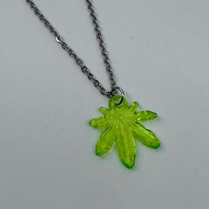 Könnte beinhalten: Halskette mit silberfarbener Kette und einem Anhänger in Form eines neongrünen Marihuanablattes. Der Anhänger hat eine durchscheinende, fünfzackige Blattform in einer hellen, auffälligen Farbe. Die Halskette ist schlicht und modern.