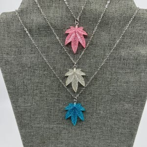May include: Three necklaces with silver chains, each featuring a glittery leaf pendant. The pendants are in pink, silver, and blue. The necklaces are displayed on a gray fabric surface.