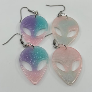 May include: Four pairs of alien-shaped earrings. Each earring is a gradient of colors, including pink, blue, and purple, with silver-colored ear wires. The earrings have a glittery, sparkly appearance.