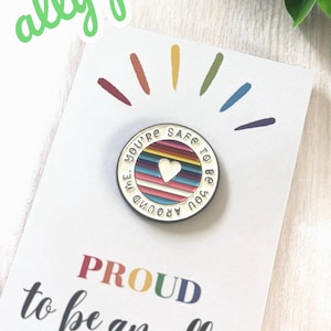 May include: A rainbow-coloured enamel pin with the text "You're safe to be you around me."  The pin has a white heart in the centre. The pin is on a white card with the text "Proud to be an ally" in rainbow colours.