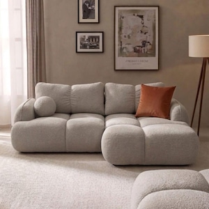 Puede incluir: Salón moderno con un sofá seccional gris claro con bordes redondeados y un puf a juego. Un cojín naranja añade un toque de color. La habitación incluye obras de arte enmarcadas y una lámpara de pie, todo sobre una alfombra de tonos neutros.