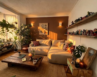 Könnte beinhalten: Ein Wohnzimmer mit einem beigen Ecksofa, einem niedrigen Couchtisch aus Holz und zwei Wandregalen, auf denen eine Sammlung kleiner Figuren steht. Der Raum enthält zahlreiche Topfpflanzen, einen gerahmten Wanddruck und eine warme Beleuchtung.