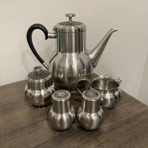 May include: A stainless steel tea set with an ornate design. The set includes a teapot, creamer, sugar bowl, and two salt and pepper shakers.