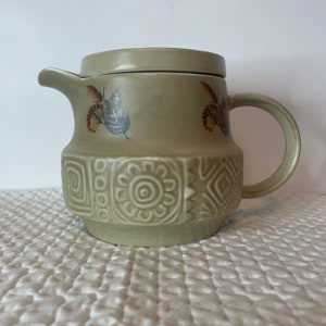 May include: A green ceramic teapot with a lid and a handle. The teapot features a geometric pattern and floral designs.