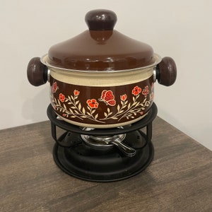 May include: A brown fondue pot with a floral design and a black metal stand. The pot has a lid and two handles. The floral design features red and white flowers and butterflies.