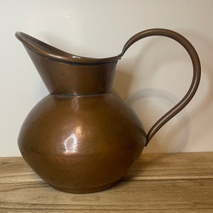 May include: A copper jug with a rounded body and a curved handle. The jug has a shiny, slightly tarnished finish.