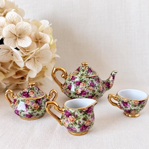 May include: A miniature porcelain tea set featuring a teapot with a lid, a lidded sugar bowl, a creamer, and a teacup. Each piece is decorated with a dense pattern of pink and yellow roses on a green background with gold trim.