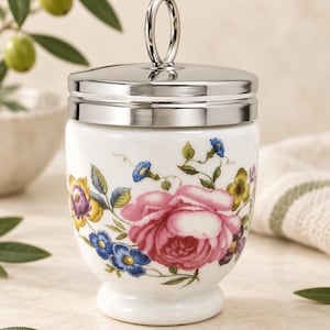 May include: A white porcelain jar with a floral pattern featuring a large pink rose, blue forget-me-nots, and yellow flowers. The jar has a polished silver-tone metal lid with a vertical ring handle on top and sits on a pedestal base.