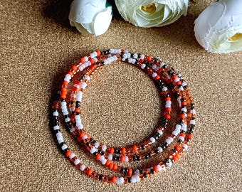 May include: A collection of beaded waist beads in orange, white, black, and gold. The beads are arranged in a circular pattern on a gold surface, with white flowers in the background.