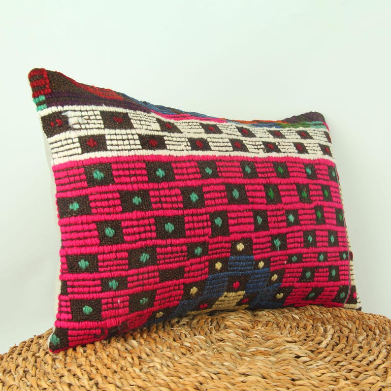 May include: A colorful rectangular pillow with a geometric pattern in shades of pink, brown, green, white, and blue. The pillow is made of woven fabric and has a fringed edge.