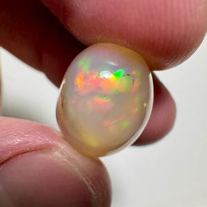 Puede incluir: Una piedra preciosa de ópalo pulida, de forma ovalada. El ópalo muestra un color base blanco lechoso con destellos internos de colores iridiscentes, incluyendo rojo, verde y amarillo. La piedra preciosa se sostiene entre dos dedos.