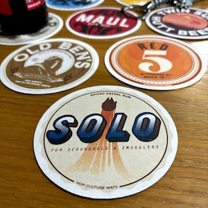 以下が含まれることがあります： 円形の茶色とベージュのコースターで、「SPICED KESSEL RUM」と「SOLO」の文字が大きく青と黒で書かれています。「FOR SCOUNDRELS & SMUGGLERS」の文字は「SOLO」の下に印刷されています。コースターには、「HOP CULTURE MATS」の文字も下部に印刷されています。