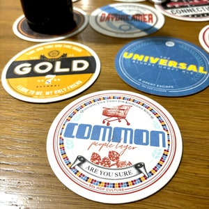 May include: A collection of colorful round coasters with various designs and text. The coasters feature a shopping cart, hops, and text such as "Common People Lager", "The Universal", "Gold", and "Daydreamer".
