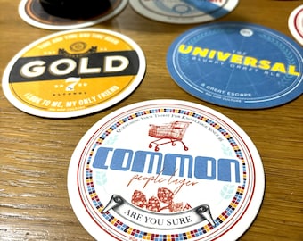 May include: A collection of colorful round coasters with various designs and text. The coasters feature a shopping cart, hops, and text such as "Common People Lager", "The Universal", "Gold", and "Daydreamer".