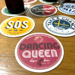 Op de afbeelding: Een verzameling ronde bierviltjes met diverse ontwerpen en tekst. Eén viltje toont de woorden "DANCING QUEEN" in een retro lettertype, een ander zegt "SOS", en weer een ander zegt "TAKE A CHANCE ON ME". De viltjes liggen op een houten oppervlak.