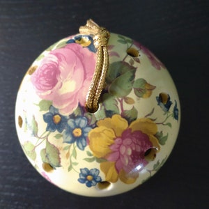 May include: A white ceramic sphere with a floral pattern in shades of pink, yellow, and green. The sphere has a gold cord attached to the top for hanging.