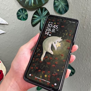 Könnte beinhalten: Ein Smartphone mit schwarzer Hülle zeigt einen digitalen Sperrbildschirm. Zu sehen sind die Uhrzeit 10:45, das Datum 26.8. und 14 Grad. Das Hintergrundbild zeigt eine weiße Katze zwischen roten Blumen und einem gelben Stern mit dem Text Zum Entsperren nach oben wischen.
