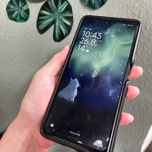 Könnte beinhalten: Ein Smartphone mit schwarzer Schutzhülle zeigt ein digitales Hintergrundbild mit einer weißen Katze in einer dunklen Landschaft unter einem Sternenhimmel mit einem leuchtenden, katzenförmigen Nebel. Das Display zeigt 10:43, 26.8 und Noch einmal.