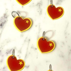 May include: Red heart-shaped keychains with a yellow border and a yellow star. Each keychain has a silver metal ring attached. The keychains are scattered on a white and gray marble surface.