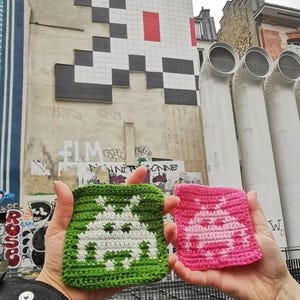 May include: Two crocheted squares, one green and one pink, each with a white pixelated alien design. They are held up in front of a building with a large, pixelated mural.