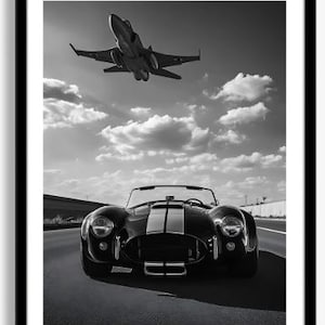 May include: A black and white framed print featuring a front view of a classic roadster with dual racing stripes on a road, positioned directly beneath a fighter jet flying through a cloudy sky. The composition captures the vehicle and aircraft.