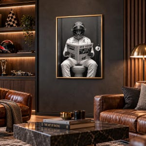May include: A framed black and white wall art print depicting a race car driver in a full white racing suit and helmet sitting on a toilet while reading a newspaper with the headline PIT STOP, mounted on a dark wall in a room.