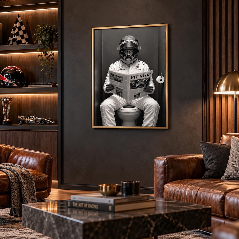 May include: A framed black and white wall art print depicting a race car driver in a full white racing suit and helmet sitting on a toilet while reading a newspaper with the headline PIT STOP, mounted on a dark wall in a room.