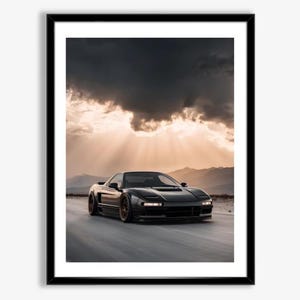 May include: Framed print of a black sports car speeding down a road. The car is the focal point, set against a backdrop of a dramatic sky with sun rays breaking through dark clouds. Mountains are visible in the distance.