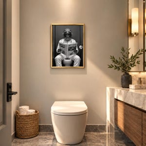 May include: A framed black and white wall art print featuring a race car driver in a full white racing suit and helmet sitting on a toilet while reading a newspaper with the headline PIT STOP. The artwork is displayed in a gold frame.