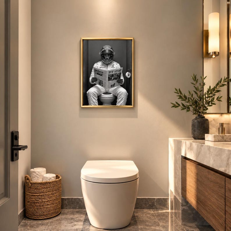 May include: A framed black and white wall art print featuring a race car driver in a full white racing suit and helmet sitting on a toilet while reading a newspaper with the headline PIT STOP. The artwork is displayed in a gold frame.