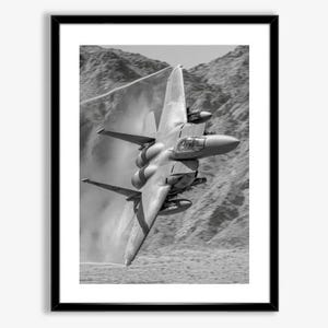 Puede incluir: Impresión enmarcada en blanco y negro de un avión de combate en vuelo. El avión está inclinado hacia abajo, con sus alas y cola visibles. El fondo muestra un paisaje montañoso con un efecto brumoso.