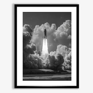 May include: A monochrome framed print depicting a space shuttle launch. The shuttle is ascending through a cloud of smoke and vapour, set against a backdrop of fluffy clouds and a dark sky. The print is framed in a black border.