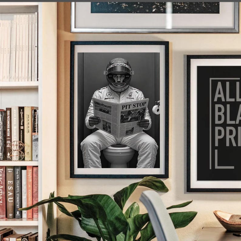 May include: A framed black and white wall art print featuring a race car driver in a full racing suit and helmet sitting on a toilet while reading a newspaper with the headline PIT STOP. The driver wears a suit with visible logos.