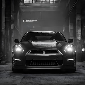 May include: A black sports car with bright headlights is parked in a dimly lit industrial setting. The car's sleek design and low profile are emphasized by the monochrome color scheme. The image captures the car's front, showcasing its grille and emblem.
