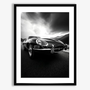 May include: A black and white photograph of a classic black Jaguar E-Type sports car driving on a road with a dramatic cloudy sky in the background.