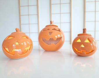 May include: Three glossy orange ceramic jack-o'-lanterns of varying sizes, each featuring carved triangular eyes, a nose, and a jagged mouth. The hollow pumpkins have a lid on top and emit a warm, soft glow from within, suggesting a light source.