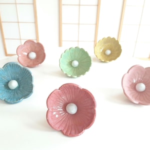 Könnte beinhalten: Sechs blumenförmige Keramikschalen in verschiedenen Pastellfarben wie Blau, Rosa, Grün und Gelb. Jede Schale hat eine glänzende Oberfläche, gewellte Blütenblattränder und eine kleine, runde, weiße Keramikperle in der Mitte der Schale.