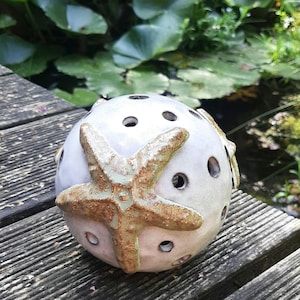 Op de afbeelding: Witte keramische bol met een zeester-ontwerp en gaten door en door. De zeester is lichtbruin met groene accenten.