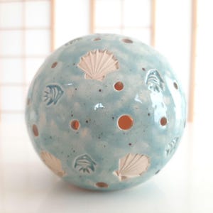 Könnte beinhalten: Ein kugelförmiger Teelichthalter aus Keramik mit hellblauer Glasur. Die Kugel ist mit weißen Muschel- und Fischmotiven verziert und hat mehrere orangefarbene Löcher. Das Gesamtdesign erinnert an ein Küstenmotiv.