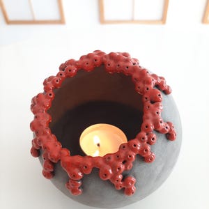 Peut inclure: Un bougeoir sphérique gris avec un bord rouge texturé. Le bougeoir contient une bougie blanche ronde allumée. Le fond est blanc avec un soupçon de cadre de fenêtre en bois.