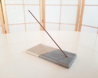 May include: A rectangular ceramic incense holder featuring a two-tone design with a speckled cream-coloured section and a solid grey section. A single thin brown incense stick is inserted into a small hole on the grey side of the holder.