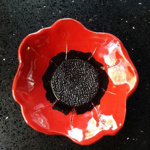 Peut inclure: Un plat en céramique en forme de coquelicot rouge avec un centre noir. Le plat est verni et a une finition brillante.