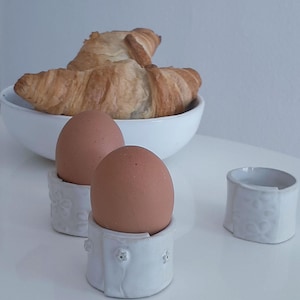 Peut inclure: Une scène de petit-déjeuner avec deux œufs bruns dans des coquetiers en céramique blanche avec des motifs floraux. Un bol blanc contient deux croissants dorés. L'ensemble est posé sur une surface blanche.