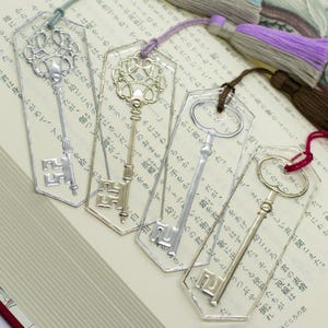 Puede incluir: Cinco marcapáginas decorativos con forma de llave y borlas de colores sobre un libro abierto. Los marcapáginas son de plástico transparente con diseños de llaves de metal ornamentados en tonos plateados y dorados. Las borlas son azules, moradas, marrones y rojas.